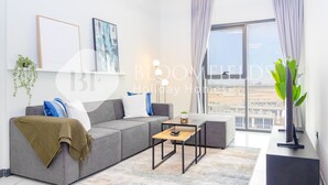 A modern living room with a grey sofa, a coffee table, and a view of a cityscape through large windows. - Bloomfields Stylish 2Br in Rukan (Dubai)