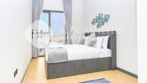 A modern bedroom with a large bed, grey curtains, a city view, and a decorative wall piece. - Bloomfields Stylish 2Br in Rukan (Dubai)