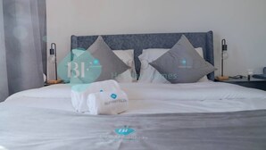 Een net opgemaakt bed met witte lakens en twee kussens, een nachtkastje met een lamp, en een wandlamp.