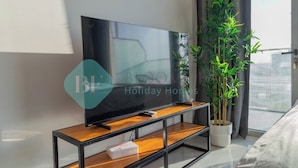 Een moderne woonkamer met een flatscreen televisie op een houten tv-meubel, een potplant en een uitzicht op de stad door grote ramen.