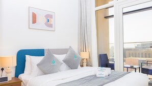 A hotel room with a bed, pillows, a bedside table with a lamp, a window with a view of buildings, and a framed picture on the wall. - Bloomfields Iconic 1BR in Oasis Masdar (Abu Dabi)