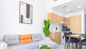 A modern living room with a grey sofa, a small table with a plant, and a dining area with a wooden table and chairs. - Bloomfields Iconic 1BR in Oasis Masdar (Abu Dabi)