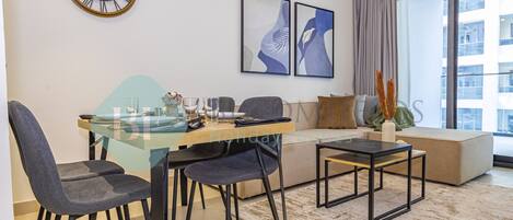 A modern living room with a glass dining table, two chairs, a sofa, a coffee table, a clock, and abstract wall art.