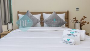 Una cama bien hecha con sábanas blancas y dos almohadas, una mesita de noche con lámpara y un reloj de pared.