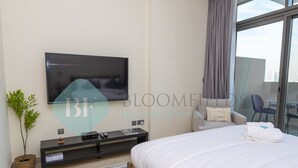 Habitación de hotel moderna con una cama grande, televisor de pantalla plana en la pared, una pequeña mesita con un control remoto, una planta en maceta y vistas de la ciudad a través de amplios ventanales.