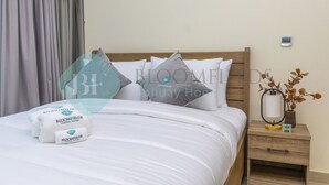 Una cama bien arreglada con sábanas blancas y un cabecero de madera, una mesita de noche con una lámpara y un pequeño jarrón con flores secas, y un termostato fijado a la pared.