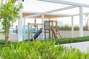 A covered outdoor area with a playground, including a slide and climbing structure, surrounded by greenery and a view of buildings in the background. - Bloomfields Stunning STU in Reflection Al Reem (Abu Dabi)