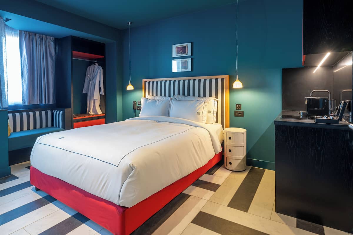 Quarto de hotel moderno com uma cama grande, mesa de cabeceira, armário e uma pequena cozinha.