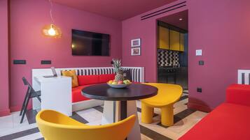 Una sala de estar moderna con un sofá rojo, sillas amarillas, una mesa redonda con fruta y un televisor en la pared.