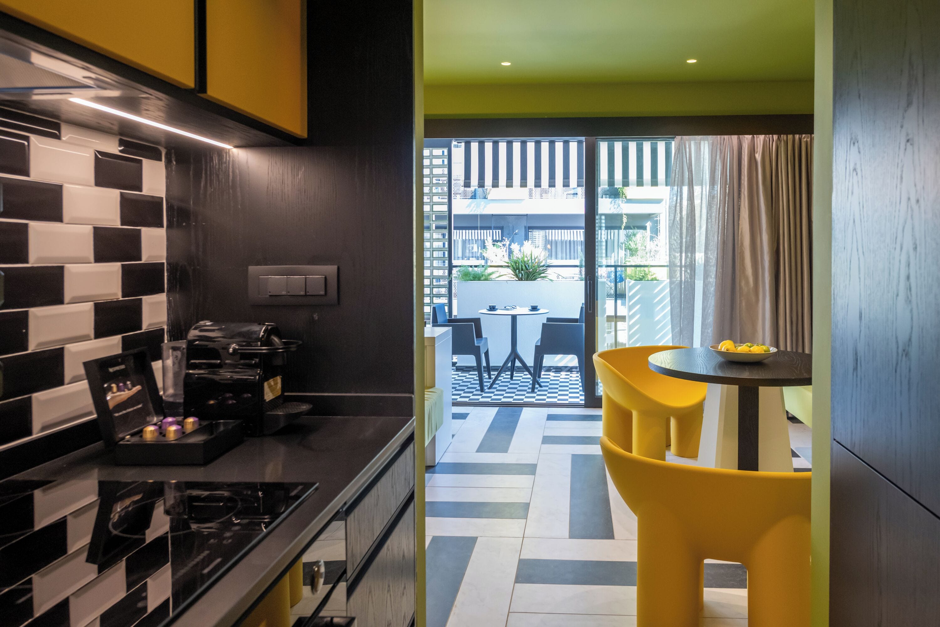 Una cocina moderna con un salpicadero a cuadros, una cafetera y un comedor con sillas amarillas y una mesa.