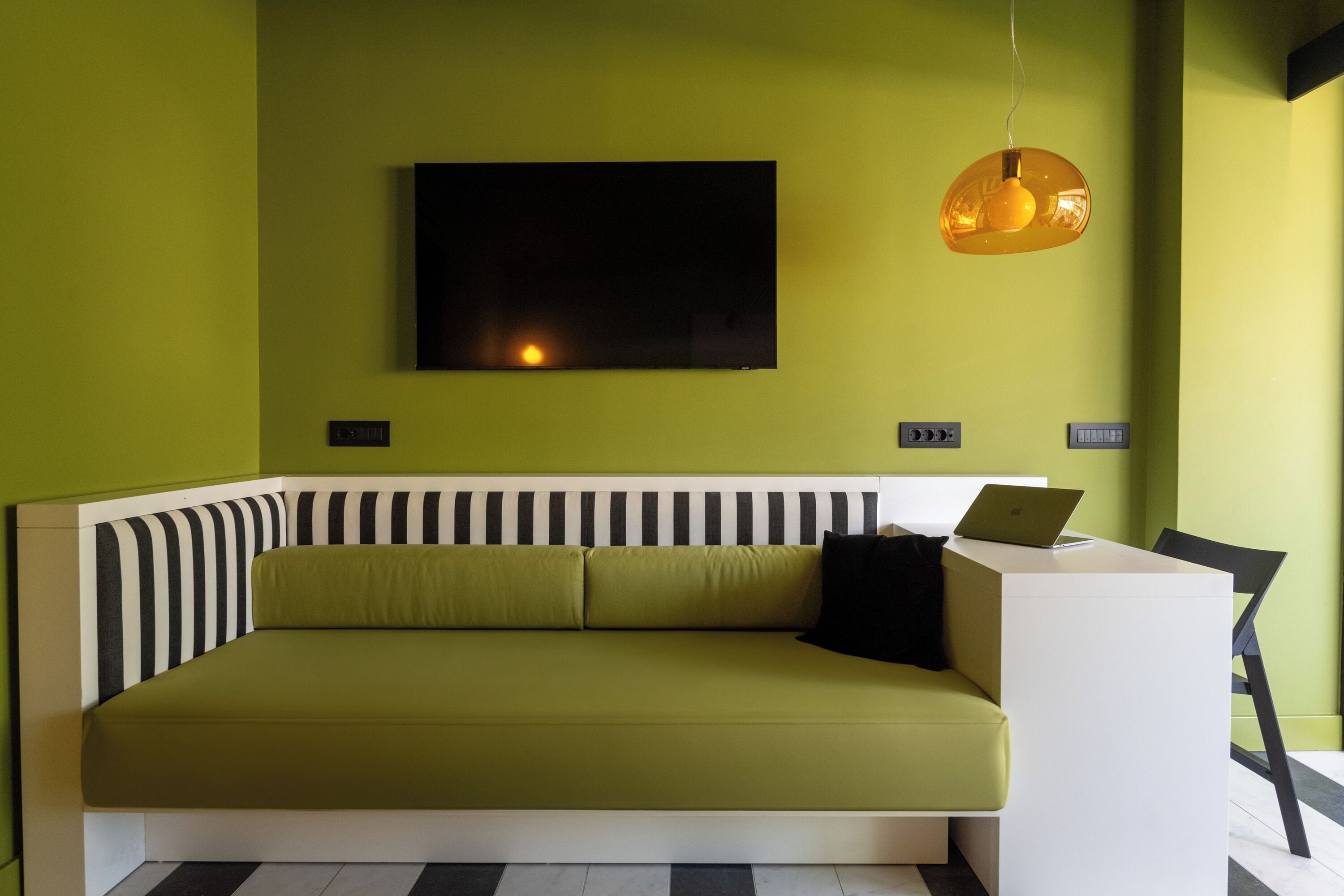 Quarto moderno com sofá verde, escrivaninha branca e cabeceira listrada em preto e branco.