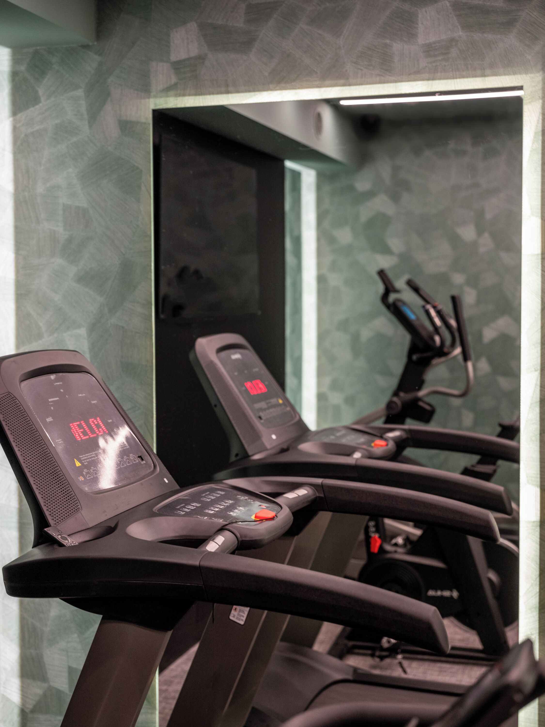 Eine Reihe stehender Ergometer mit digitalen Anzeigen vor einer Wand mit Muster.