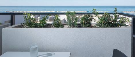 En balkon med et bord, en stol og et glas vand. Udsigten viser en strand og havet.