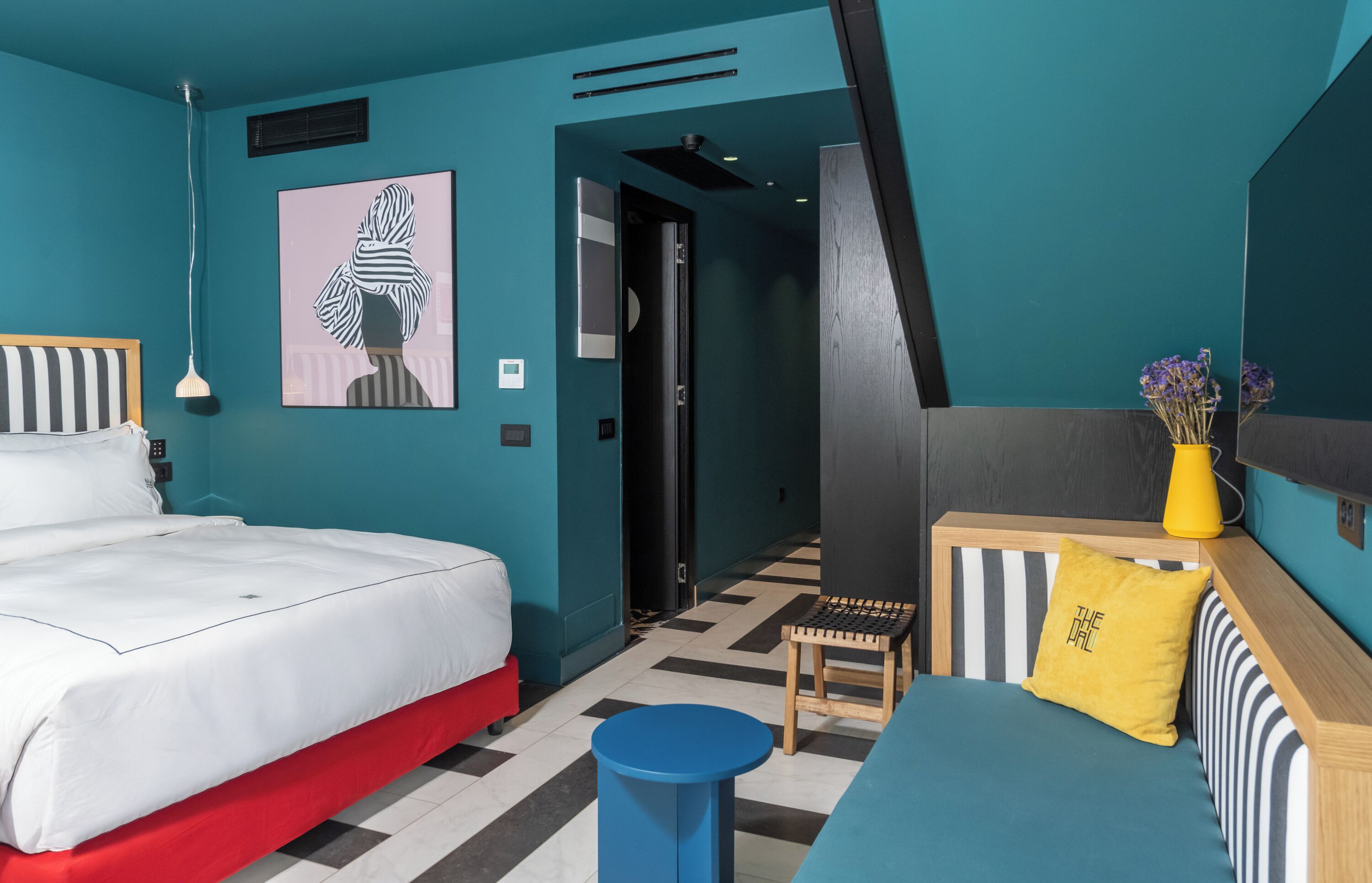 A modern hotel room with a teal color scheme, a large bed, a small table, a bench, and a bedside table with a vase of flowers.
