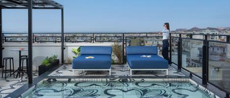 Eine Dachterrasse mit zwei blauen Liegestühlen, einem kleinen Tisch und einem Glasgeländer mit Blick auf eine Stadtlandschaft.