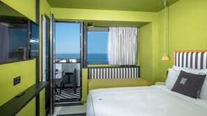 A hotel room with a bed, a desk, a chair, a window with curtains, and a view of the sea. - The Pal Living in a Concept (Heraklion)