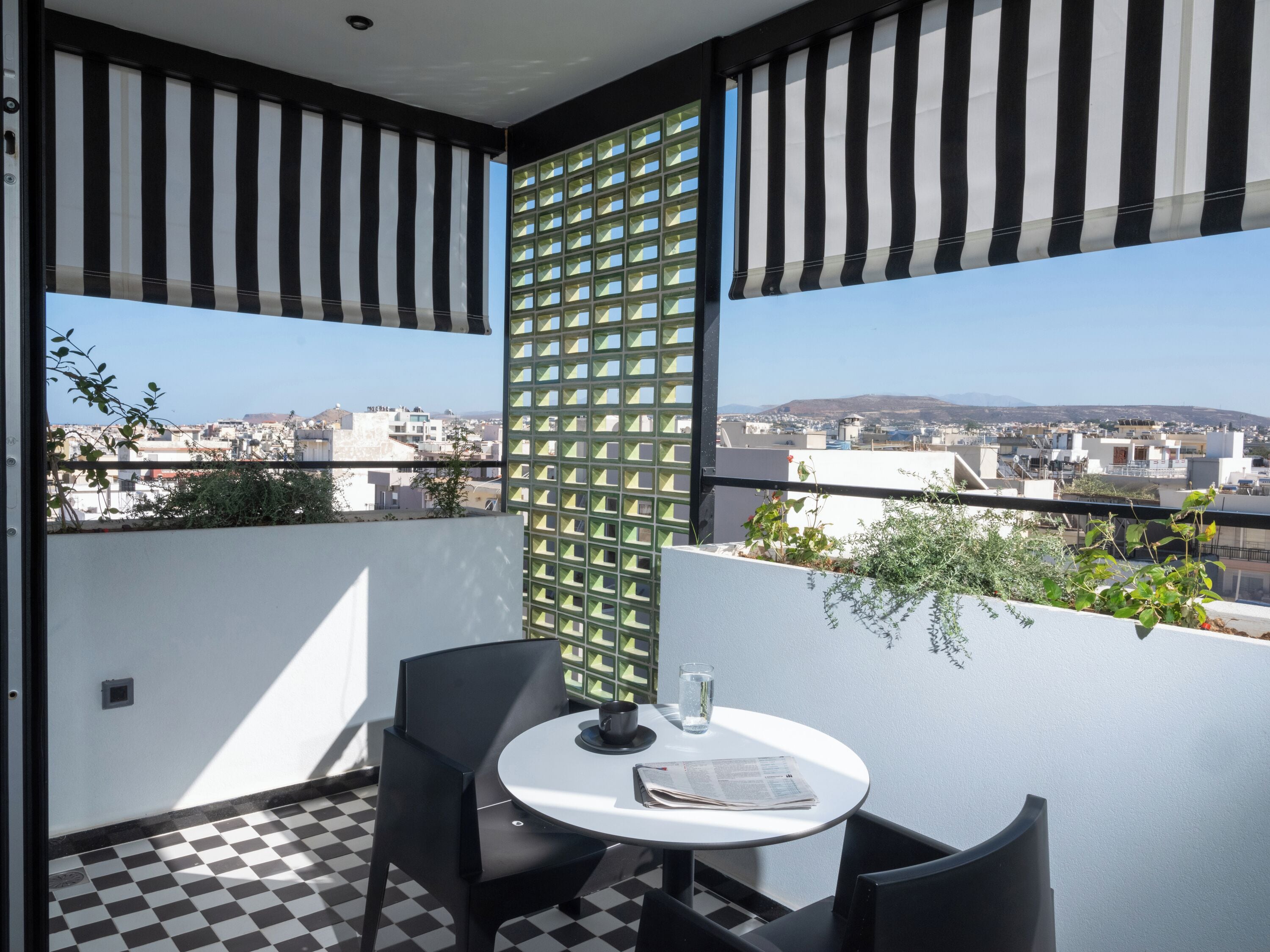 A balcony with a checkered floor, a round table with chairs, a newspaper, a glass of water, and a view of a cityscape with buildings and mountains in the distance.