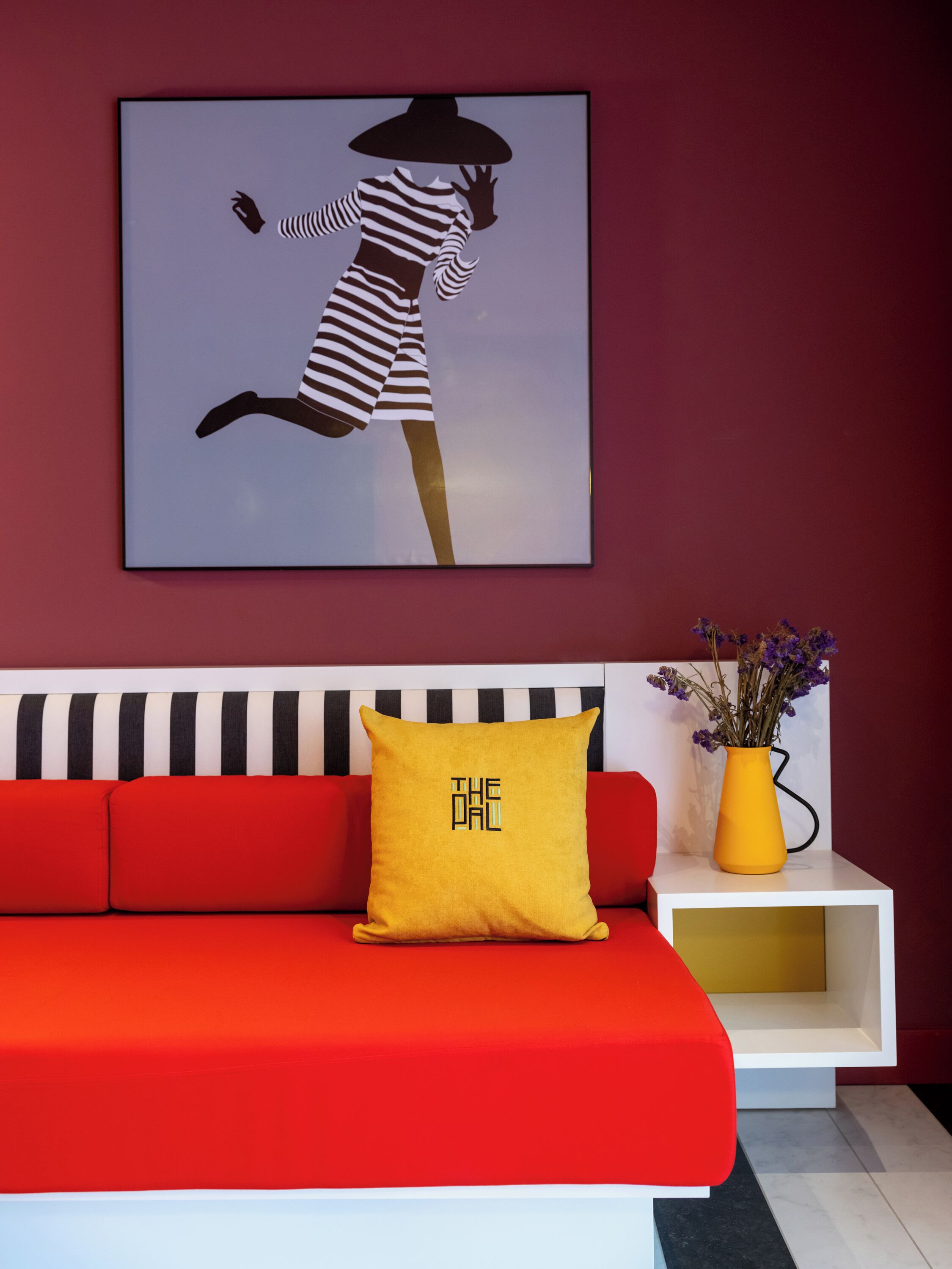 A modern living room with a red sofa, a yellow pillow with text, a black and white striped cushion, a vase with flowers, and a framed artwork on the wall.