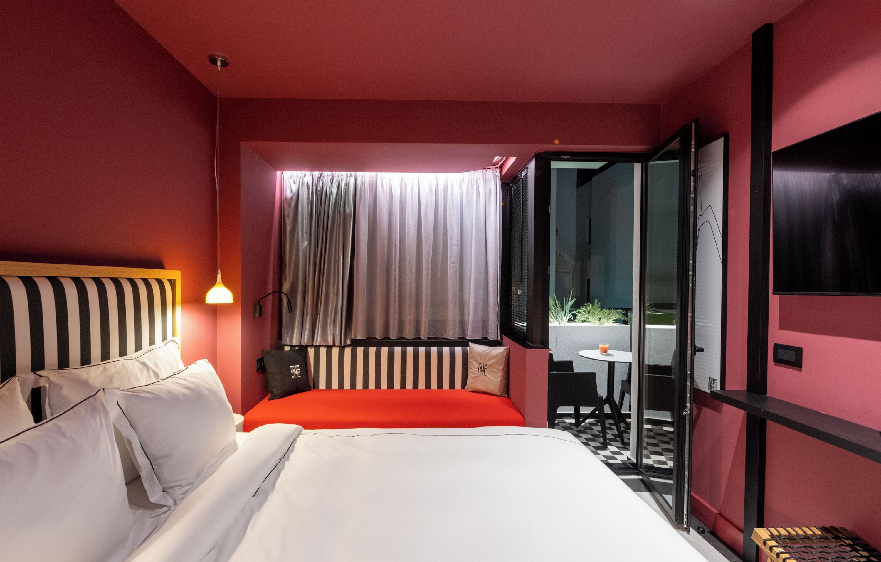 A hotel room with a large bed, a red accent wall, a view of a balcony, and a modern design.