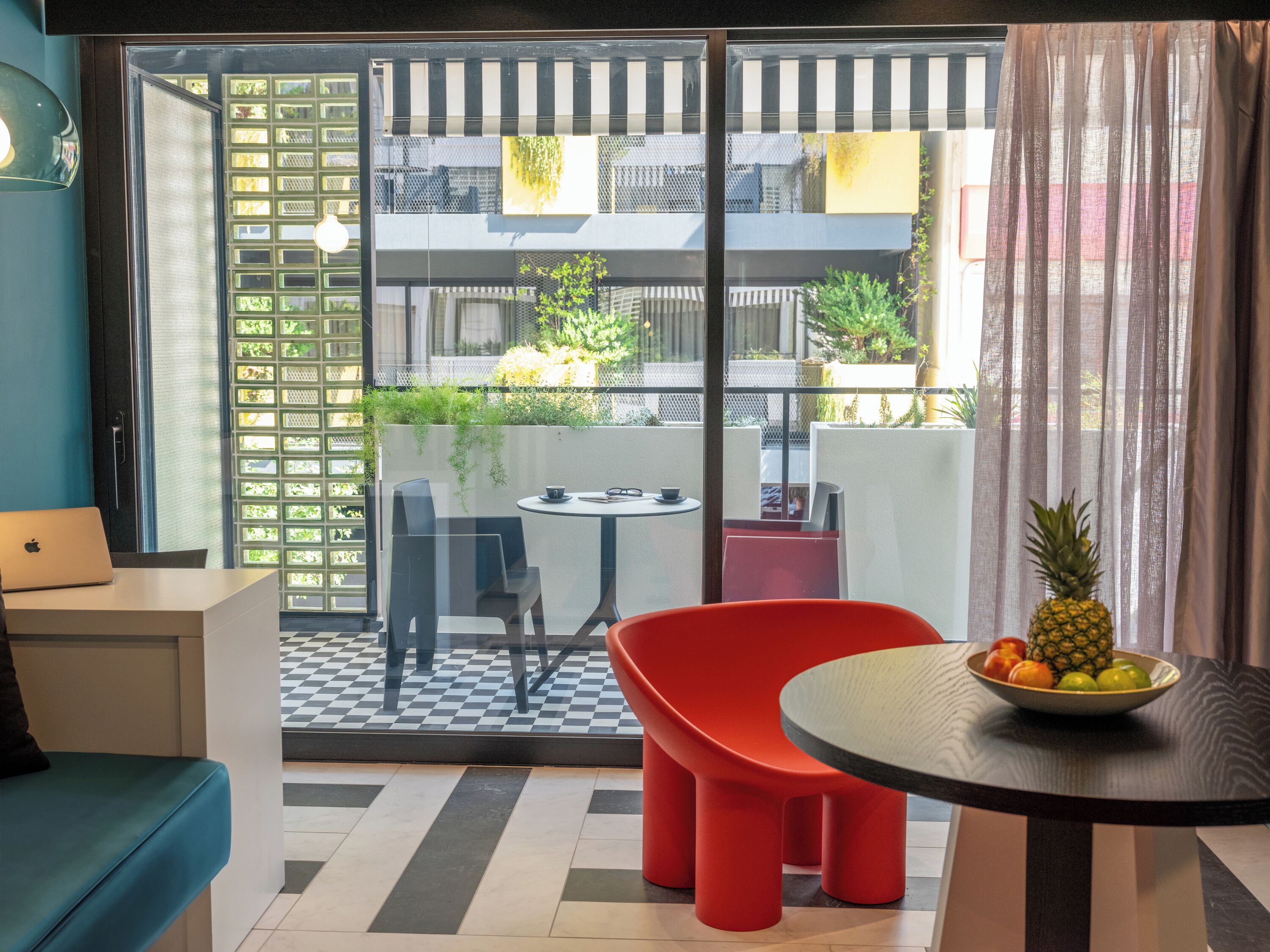 A modern room with a checkered floor, a blue sofa, a red chair, a small table with a fruit bowl, and a balcony with a view of the city.
