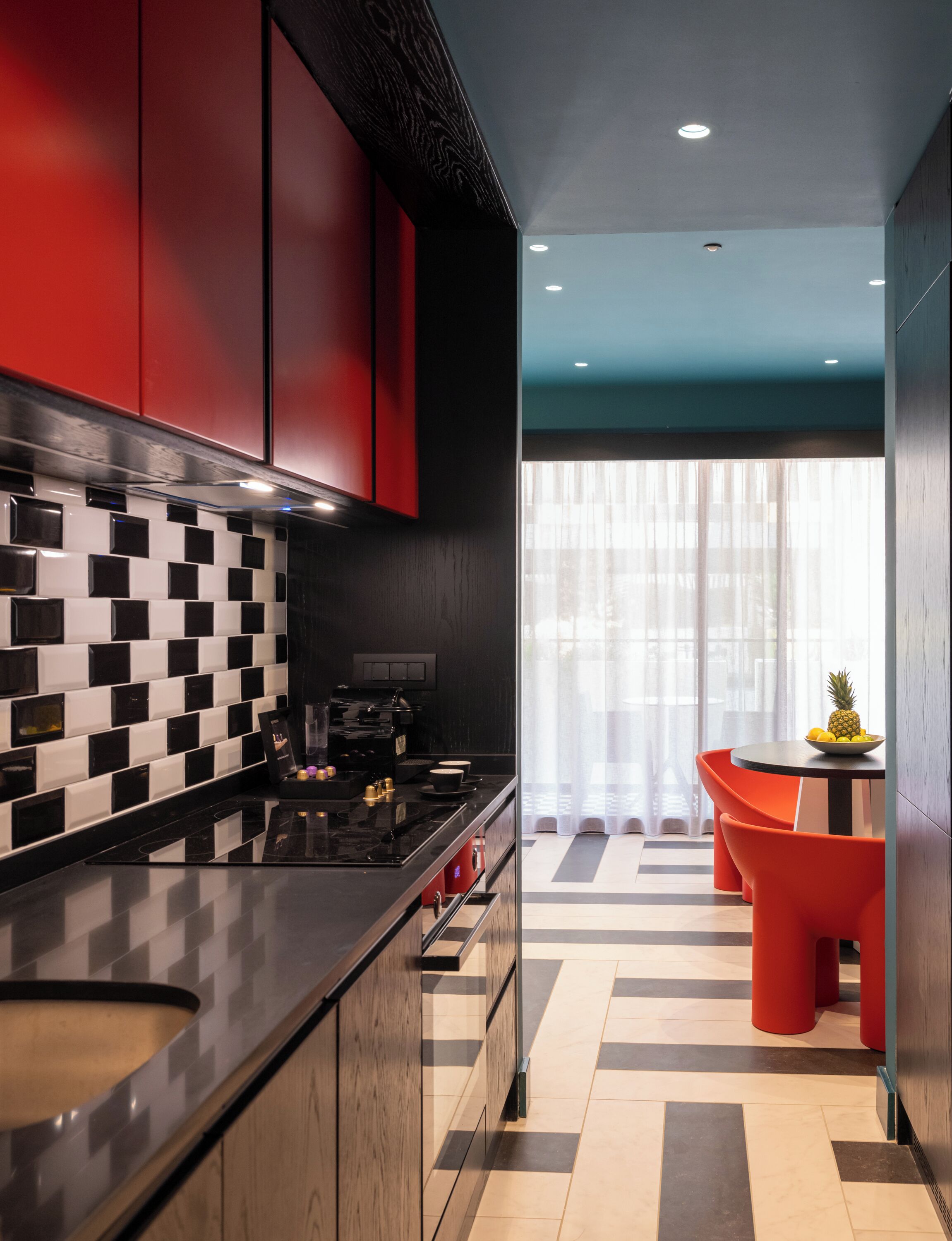A modern kitchen with black and white checkered backsplash, red cabinets, and a black countertop. There is a dining area with orange chairs and a table, and a window with sheer curtains.