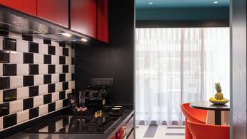 Cocina moderna con salpicadero a cuadros blancos y negros, armarios rojos y encimera negra. Hay una zona de comedor con sillas naranjas y una mesa, y una ventana con cortinas translúcidas.