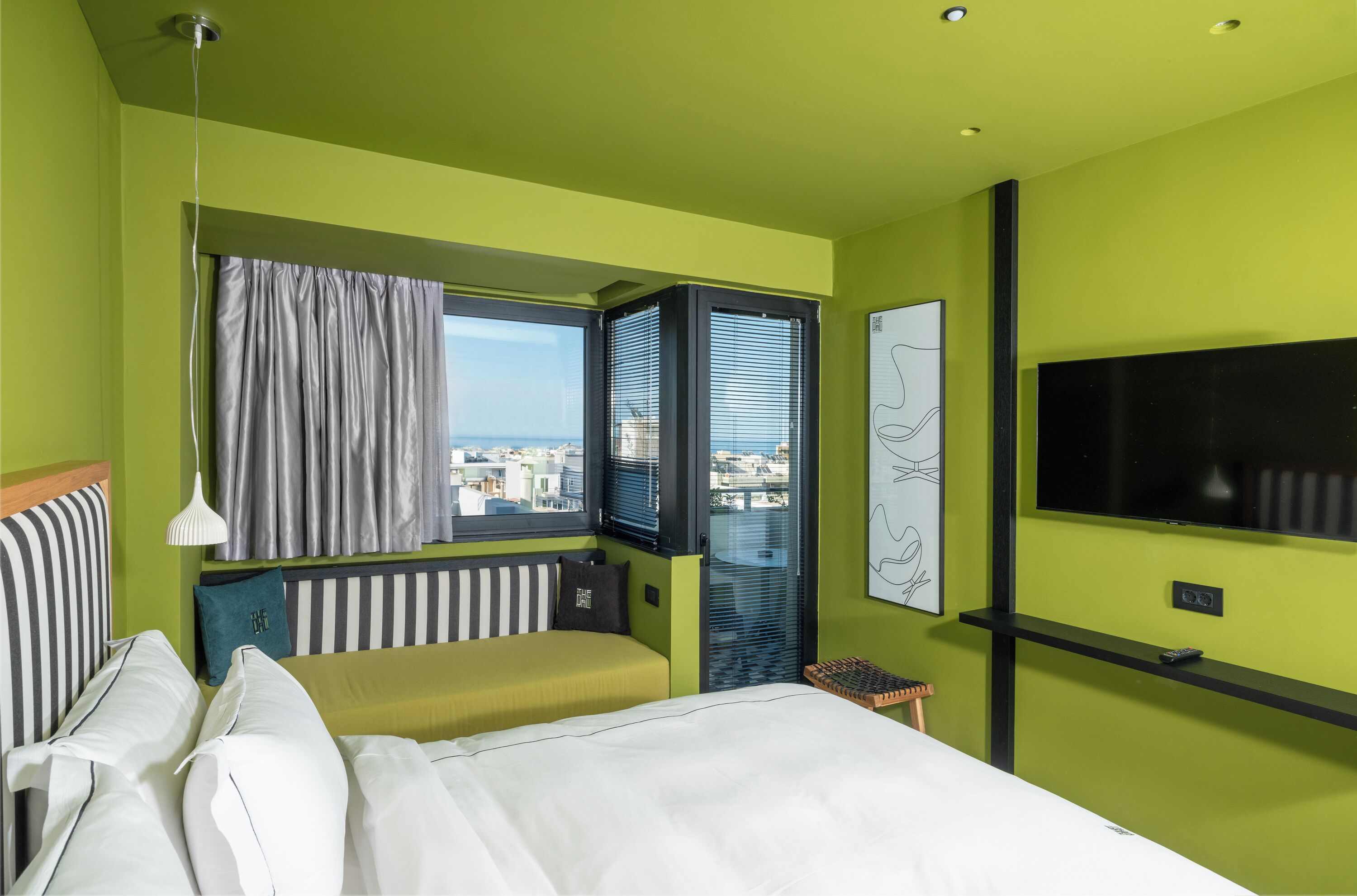A modern hotel room with a green wall, a large bed, a window with blinds, a TV, and a city view.