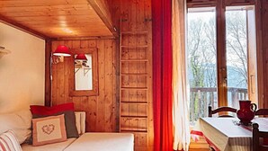 A cozy room with a wooden interior, a bed with red pillows, a table with a red pitcher, and a view of trees through a window. - 6-person apartment (PASSY)