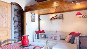 A cozy room with a wooden ceiling, a sofa with red cushions, a table with a red pitcher, and a wall with a shelf and a lamp. - 6-person apartment (PASSY)