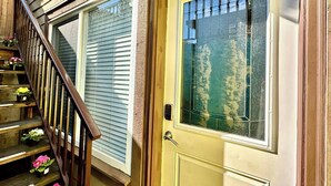 Una escalera de madera con plantas en macetas, una puerta amarilla con un ventanal y un ventano con persianas.