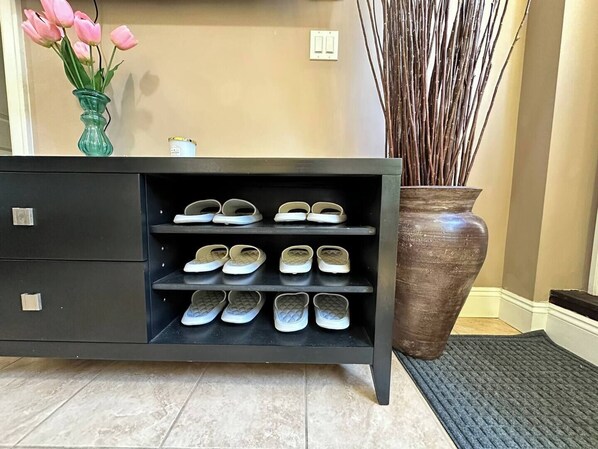 Un módulo de almacenamiento negro con varias estanterías que contienen zapatos, un jarrón grande con plantas altas y un interruptor de pared.