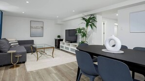 A modern living room with a grey sofa, a glass coffee table, a TV on a white cabinet, and a dining area with a black table and chairs. - Cozy Basement suite Near UBC, Kits, and DT (Vancouver)