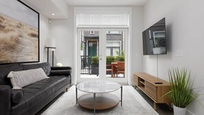 A modern living room with a black leather sofa, a glass coffee table, a wooden side table, and a TV mounted on the wall.