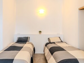 Une petite chambre propre, avec deux lits simples, une petite plante posée sur une étagère et un luminaire fixé au mur.