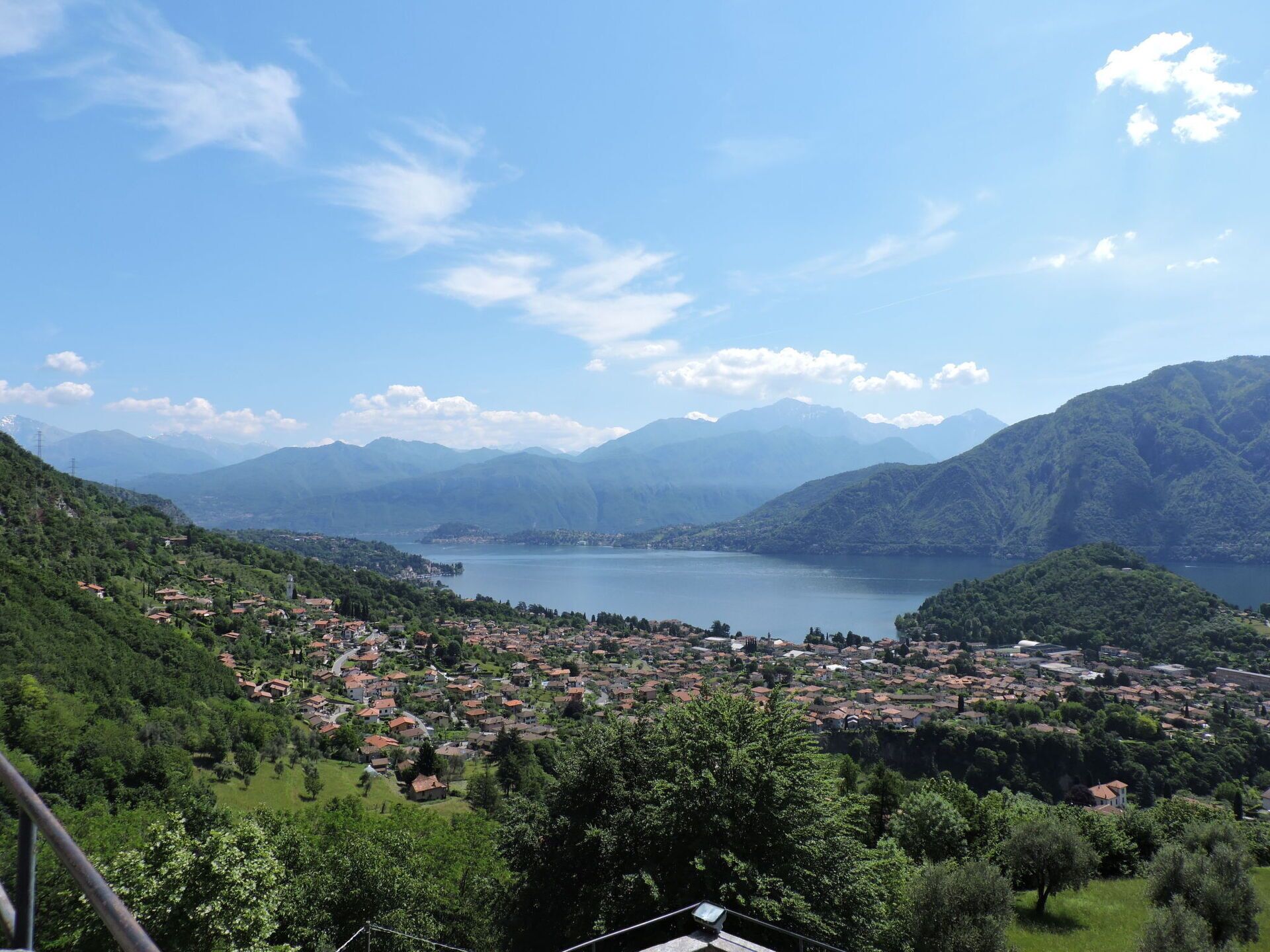 Een panoramisch uitzicht op een stad aan een meer, met een dichte bebouwing, omringd door groene heuvels en bergen op de achtergrond.