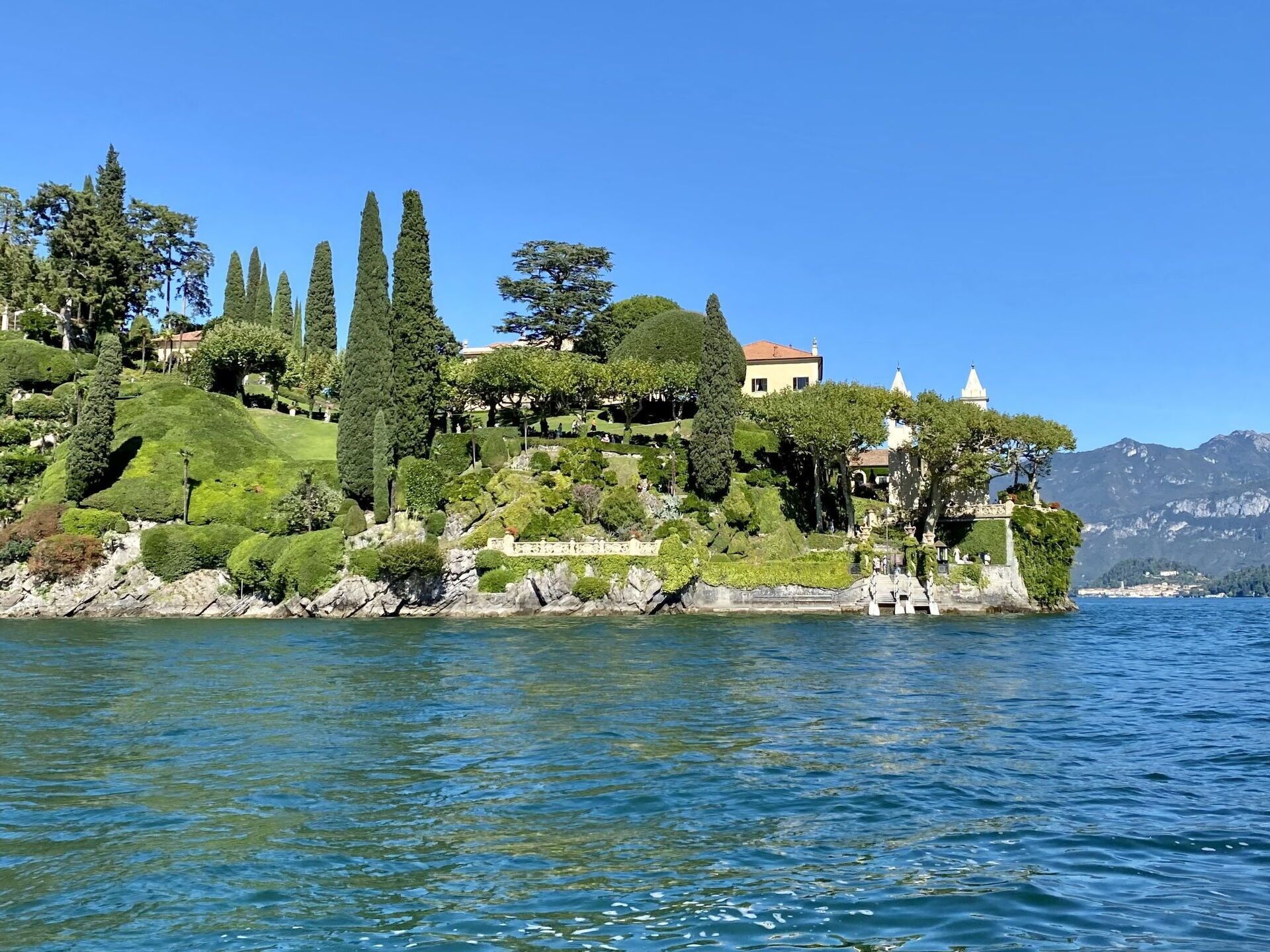 A coastal landscape with a hill featuring manicured gardens, a white building with a dome, and a statue.