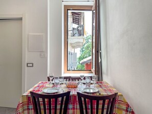 Un coin repas avec une table dressée pour deux personnes, une fenêtre donnant sur un balcon et une porte située à gauche.