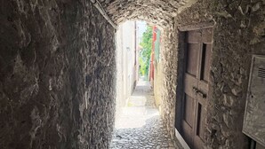 Ein schmaler Steinkorridor mit einem Kopfsteinpflasterweg, der zu einer Holztür und einem Fenster mit einem Metallgitter führt.