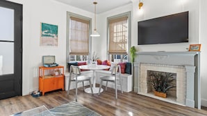A modern living room with a fireplace, a television mounted above it, and a dining area with a table and chairs. - Old Spanish House Vivian Suite A. Sophisticated Retreat, Beach, Fire Pit, Downtown, Golf Cart (Biloxi)