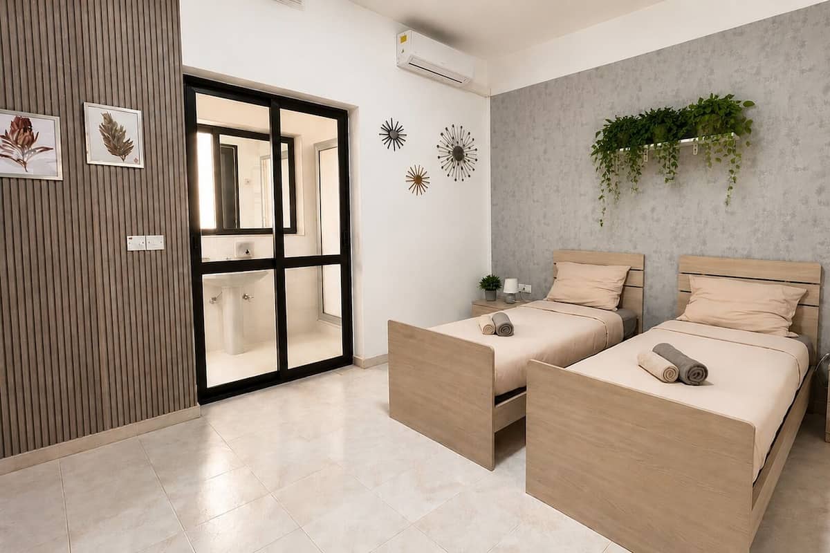 A modern bedroom with two beds, a large sliding glass door, a grey textured wall, and a small plant hanging on the wall.