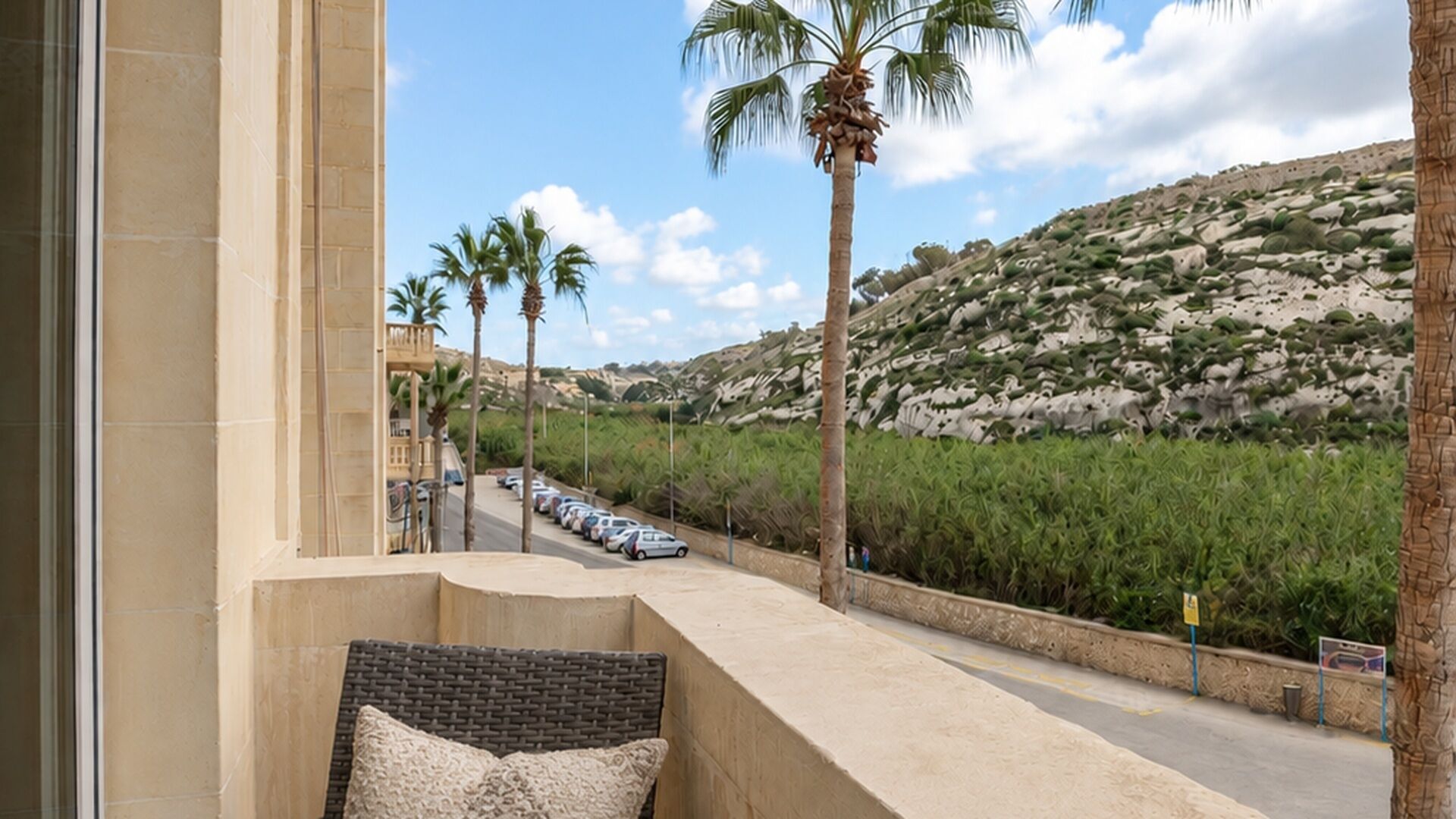 A balcony with a view of a road lined with parked cars, palm trees, and a hilly landscape.