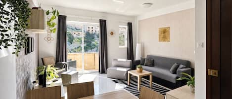 A modern living room with a sofa, coffee table, and chairs, a balcony with curtains, and a view of buildings.