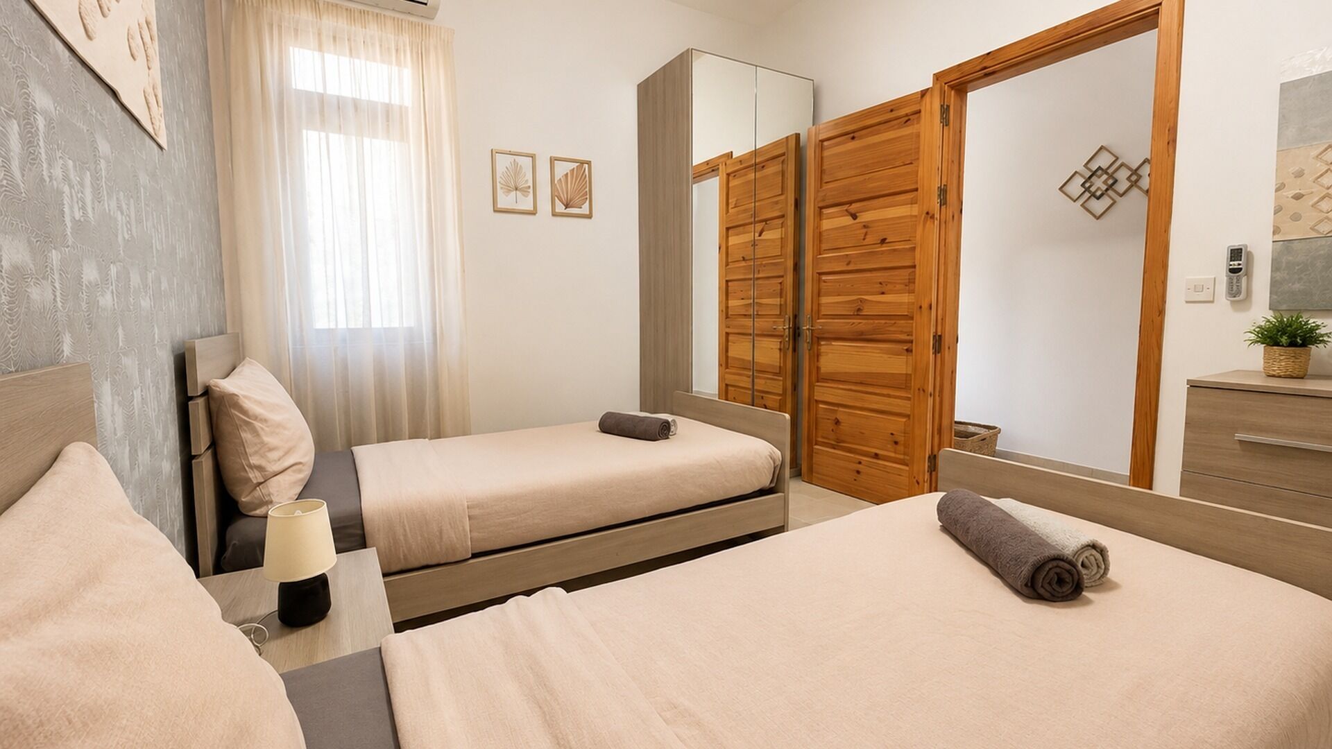 A hotel room with two beds, a wooden wardrobe, a mirror, a bedside table with a lamp, and a small plant on the dresser.