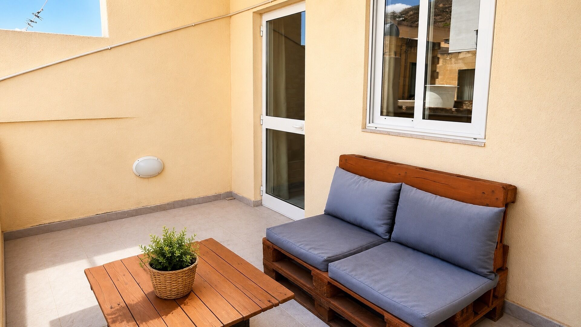 A balcony with a wooden table, a wicker plant pot, and a blue cushioned bench.