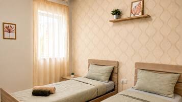 Une chambre avec deux lits, une fenêtre avec des rideaux légers, une étagère fixée au mur ornée d’une plante et d’un tableau, et un mur à motifs.