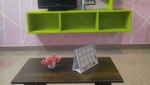 A room with a green shelving unit against a pink wall, a flat-screen TV, a small table with a calendar, and a vase with flowers. - 1BHK Spacious stay near Ramana Ashram in Tiruvannamalai (Tiruvannamalai)