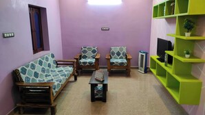 A living room with a sofa, two armchairs, a coffee table, a TV, and a green shelving unit. - 1BHK Spacious stay near Ramana Ashram in Tiruvannamalai (Tiruvannamalai)