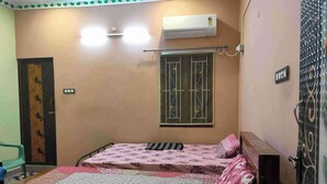A small, single-room bedroom with a bed, an air conditioning unit, a window with bars, and a door with a decorative panel. - 1BHK Spacious stay near Ramana Ashram in Tiruvannamalai (Tiruvannamalai)