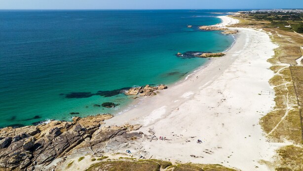Luftaufnahme eines Strandes mit klarem türkisblauem Wasser, weißem Sandstrand und Felsformationen.