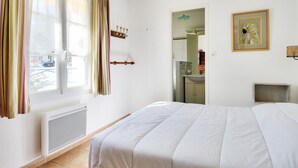 A bedroom with a large bed, a radiator, a window with curtains, a door, a kitchen area, and a framed picture on the wall. - House for 7 people 900m from the beach (Barbâtre)